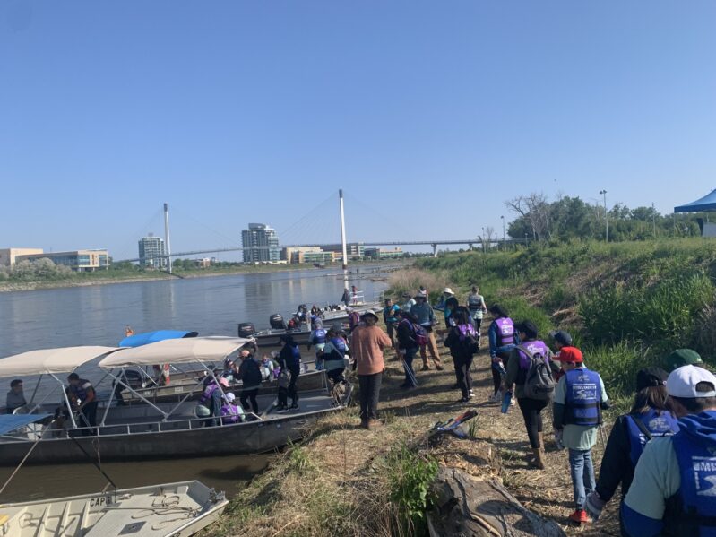 A group of people walk together along a riverside path with buildings visible in the distance.